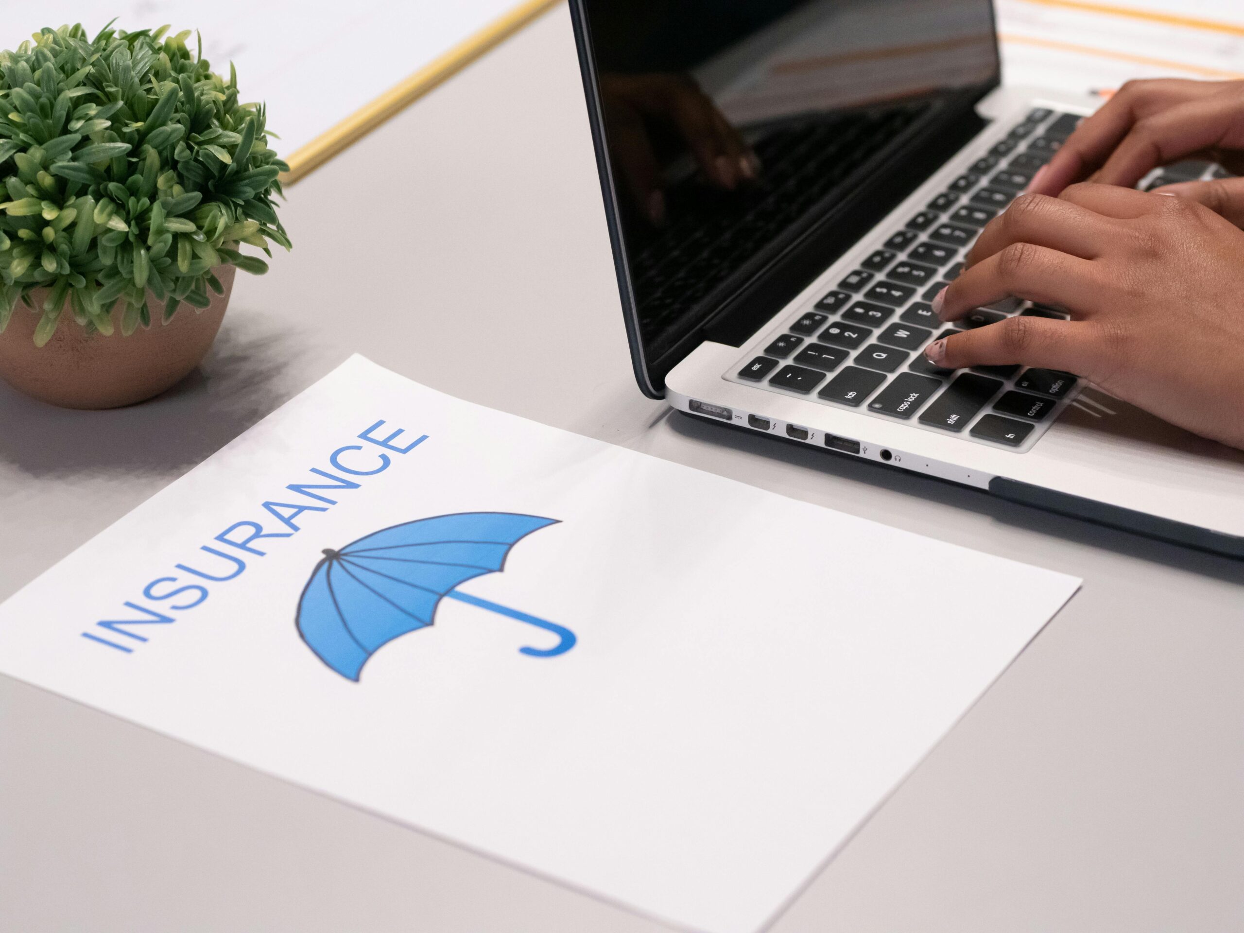 A person types on a laptop next to a paper that reads INSURANCE with a blue umbrella icon.