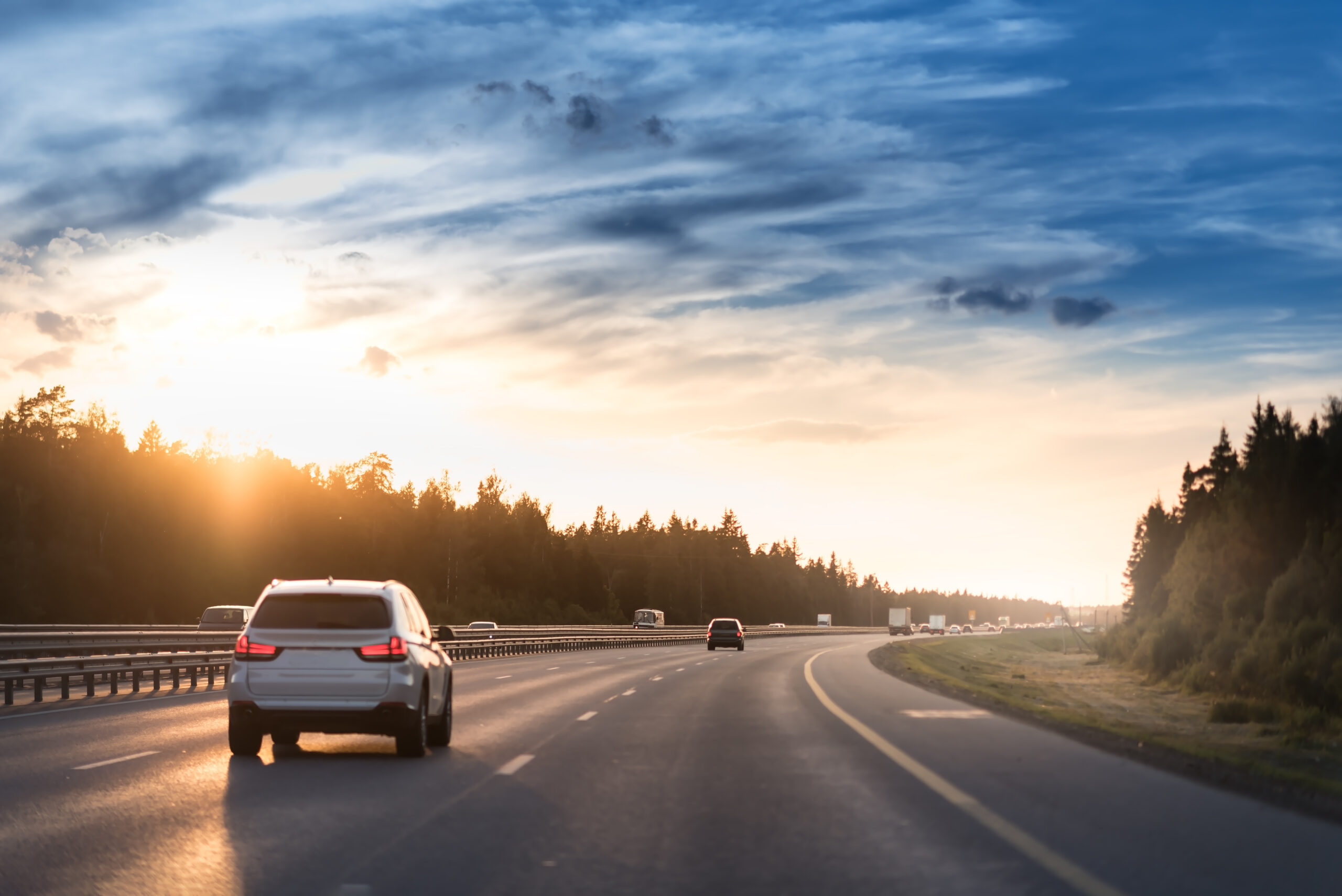 A white SUV drives on a multi-lane highway at sunset, with silhouetted trees along the roadside and a dramatic blue and orange cloudy sky above.