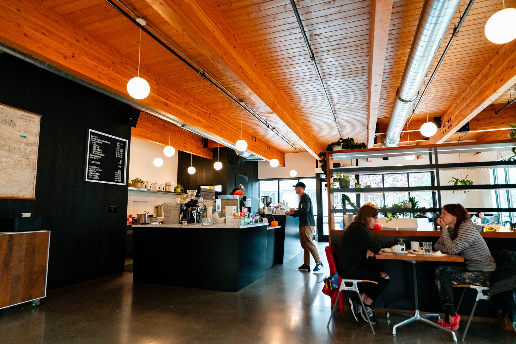 Interior view of a modern coffee shop with wooden beam ceilings, spherical pendant lights, a dark counter bar, and customers seated at a table.