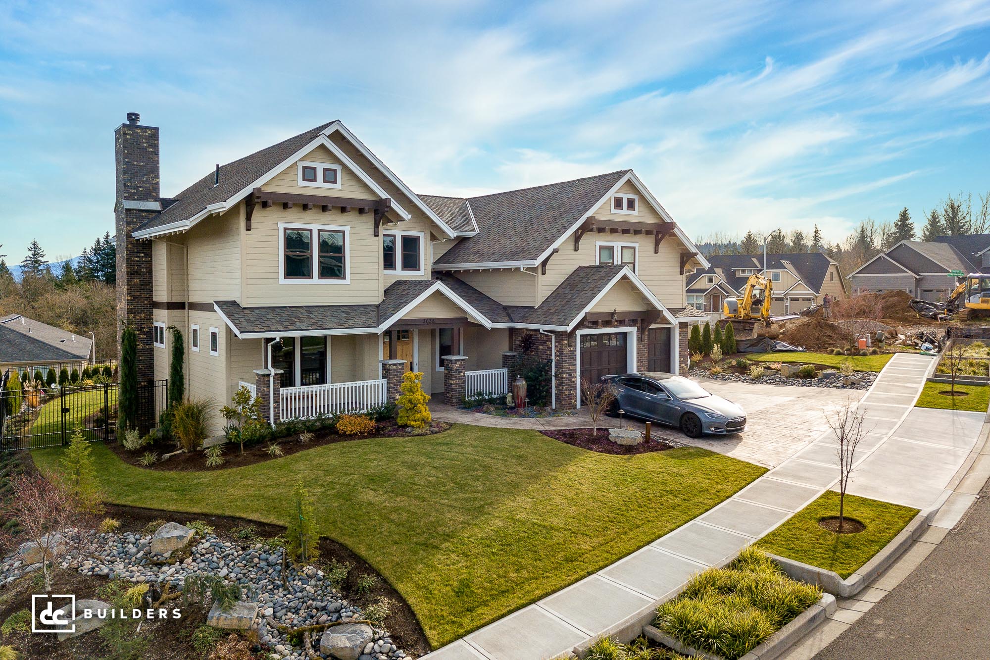 Two-story Craftsman-style house with light yellow siding, dark shingled roofs, stone accents, a chimney, and a driveway with a car.